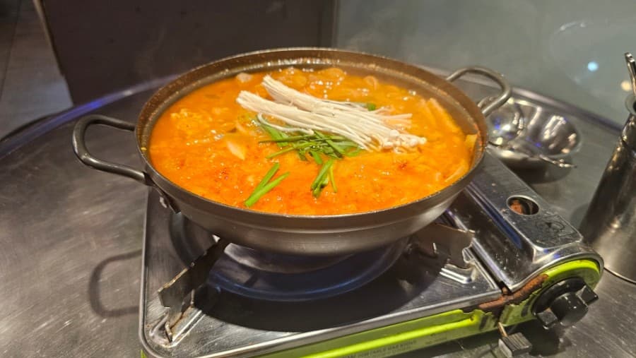 부산 북구 백양대로 1065에 위치한 노포 구남포차의 김치찌개. 팽이버섯과 부추가 올라간 매콤한 국물이 휴대용 버너 위 냄비에서 보글보글 끓고 있습니다. 혼술이나 혼밥하기 좋은 정겨운 분위기가 느껴지는 스테인리스 테이블 위 안주 사진입니다.