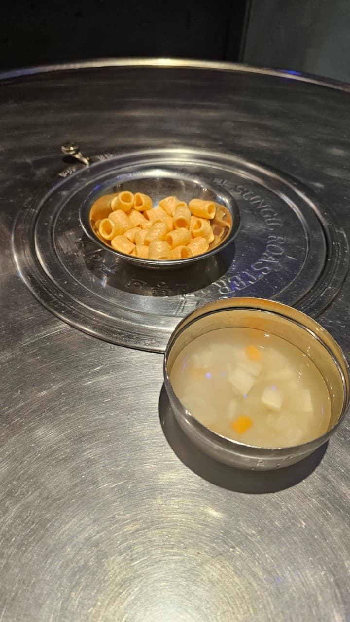 부산 북구 백양대로 구남포차의 정겨운 노포 분위기. 원형 철제 테이블 위에 기본 안주인 마카로니 뻥튀기와 시원한 동치미가 스텐 그릇에 담겨 있습니다. 혼술이나 혼밥을 즐기기 좋은 아늑하고 소박한 감성이 느껴지는 풍경입니다.