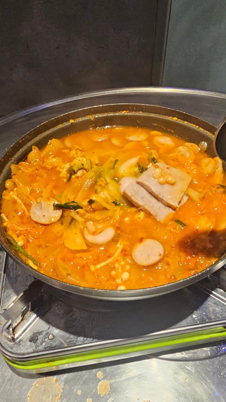부산 북구 백양대로 1065 구남포차의 보글보글 끓는 부대찌개. 햄, 소시지, 김치가 듬뿍 담긴 얼큰한 국물 요리로 노포에서 혼술이나 혼밥 하기에 제격입니다. 휴대용 가스레인지 위 냄비에 담긴 따뜻하고 먹음직스러운 안주 이미지입니다.