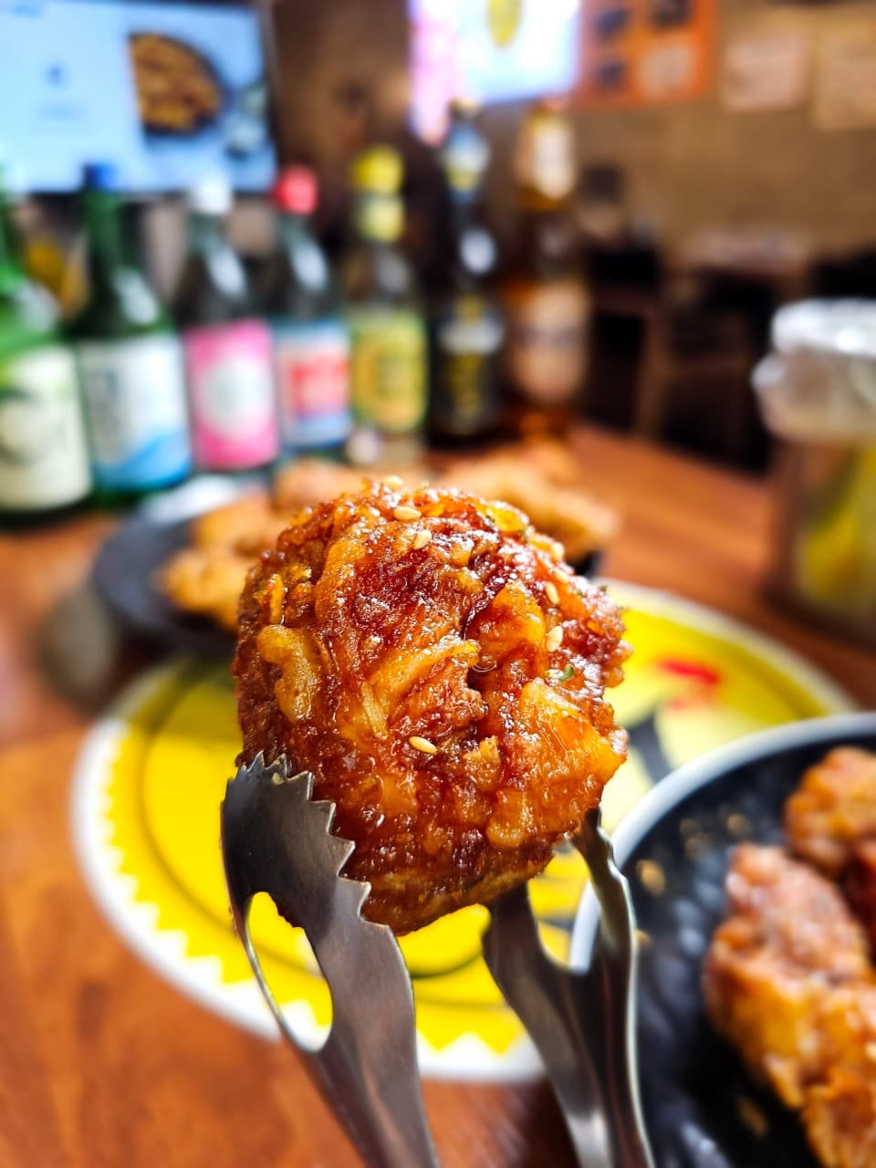 집게로 집어 올린 참깨가 뿌려진 양념 치킨과 배경의 목재 테이블 위 일렬로 늘어선 여러 종류의 주류 병들