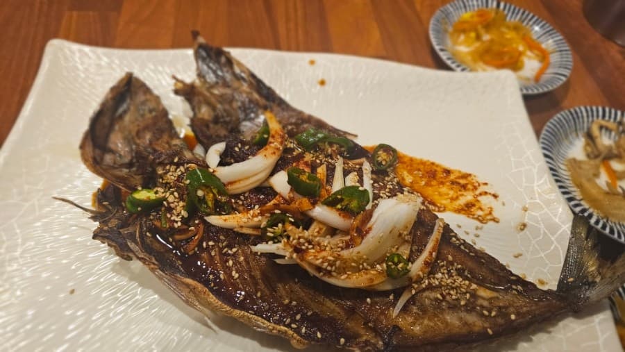 나무 식탁 위 흰 접시에 담긴 양념 생선구이와 그 위에 얹어진 양파, 청고추 고명 및 작은 찬그릇들