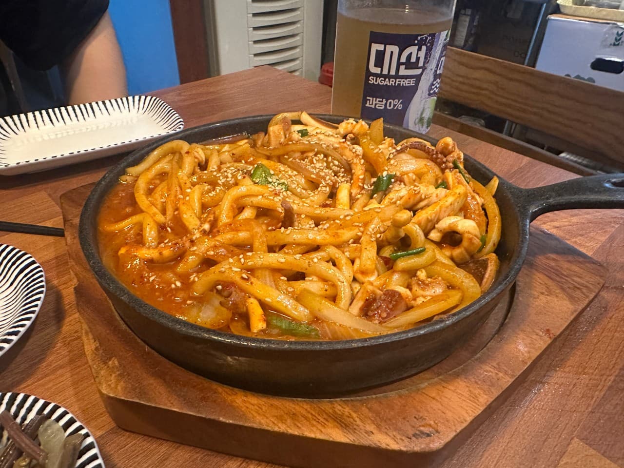 나무 테이블 위 무쇠 팬에 담긴 빨간 해물 볶음 우동과 소주병 그리고 줄무늬 앞접시