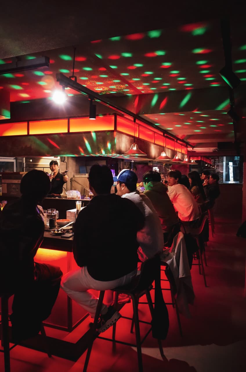 붉은 조명의 바 카운터에 나란히 앉은 손님들과 천장에 비친 원형 조명 패턴 및 오픈 주방 내부