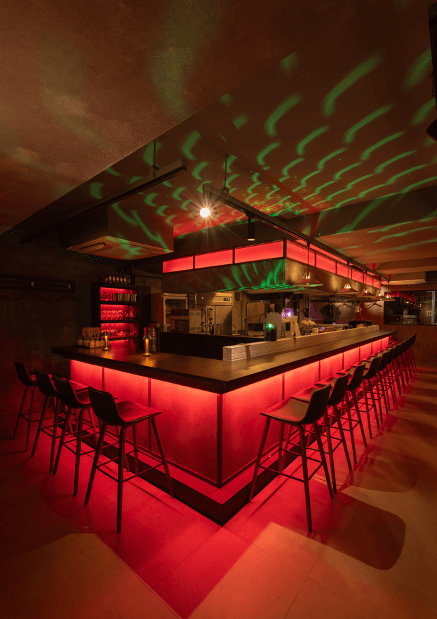 붉은 하단 조명이 들어오는 긴 바 테이블과 일렬로 놓인 1인용 높은 의자들 및 천장의 초록색 조명