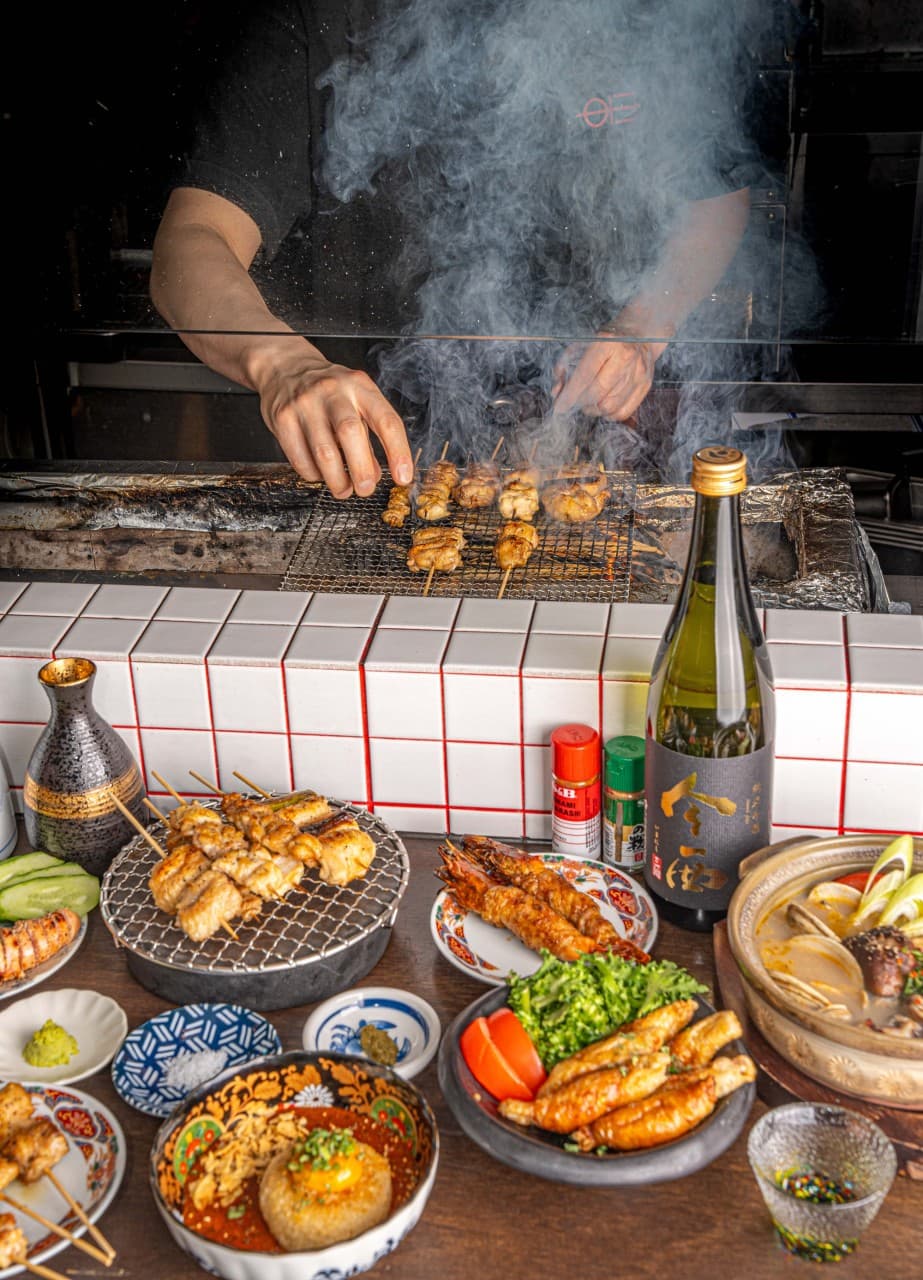 숯불 연기 속에서 꼬치를 굽는 모습과 흰 타일 카운터 위 사케 병, 닭꼬치, 나베 요리가 차려진 1인 바 테이블