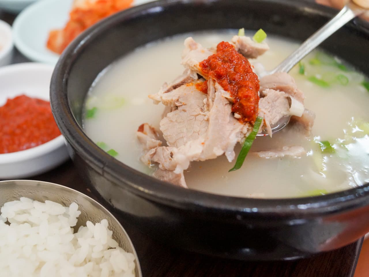 뽀얀 국물이 담긴 뚝배기에서 양념장을 얹은 수육을 숟가락으로 들어 올린 모습과 흰 쌀밥이 차려진 1인 상차림