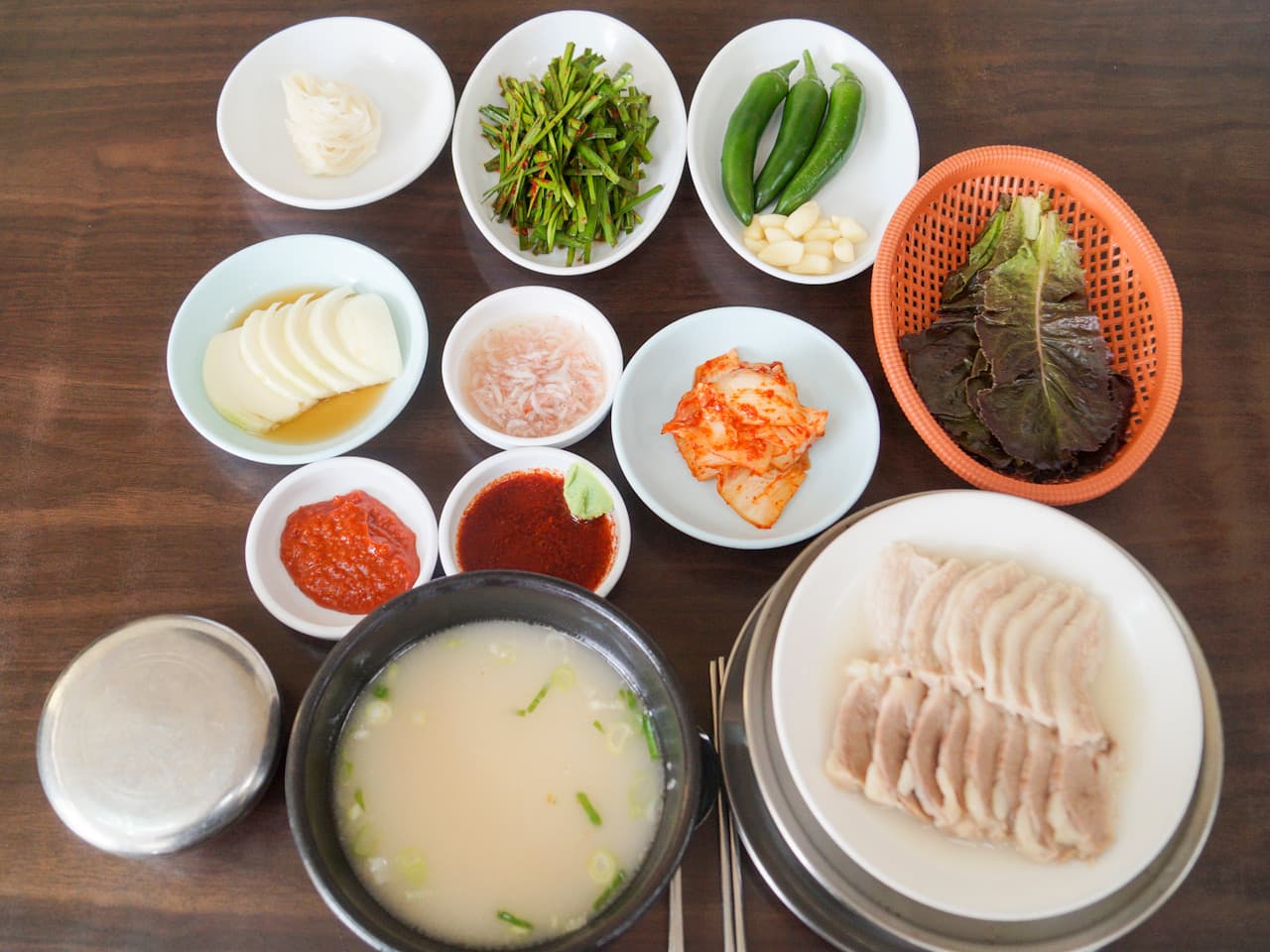 뽀얀 국물의 뚝배기와 수육, 부추무침, 쌈 채소 등 여러 반찬이 놓인 목제 테이블 위 1인 상차림