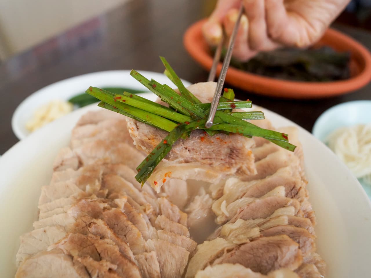 가지런히 정렬된 수육 중 한 점을 젓가락으로 집어 부추를 곁들인 모습과 옆에 놓인 소면