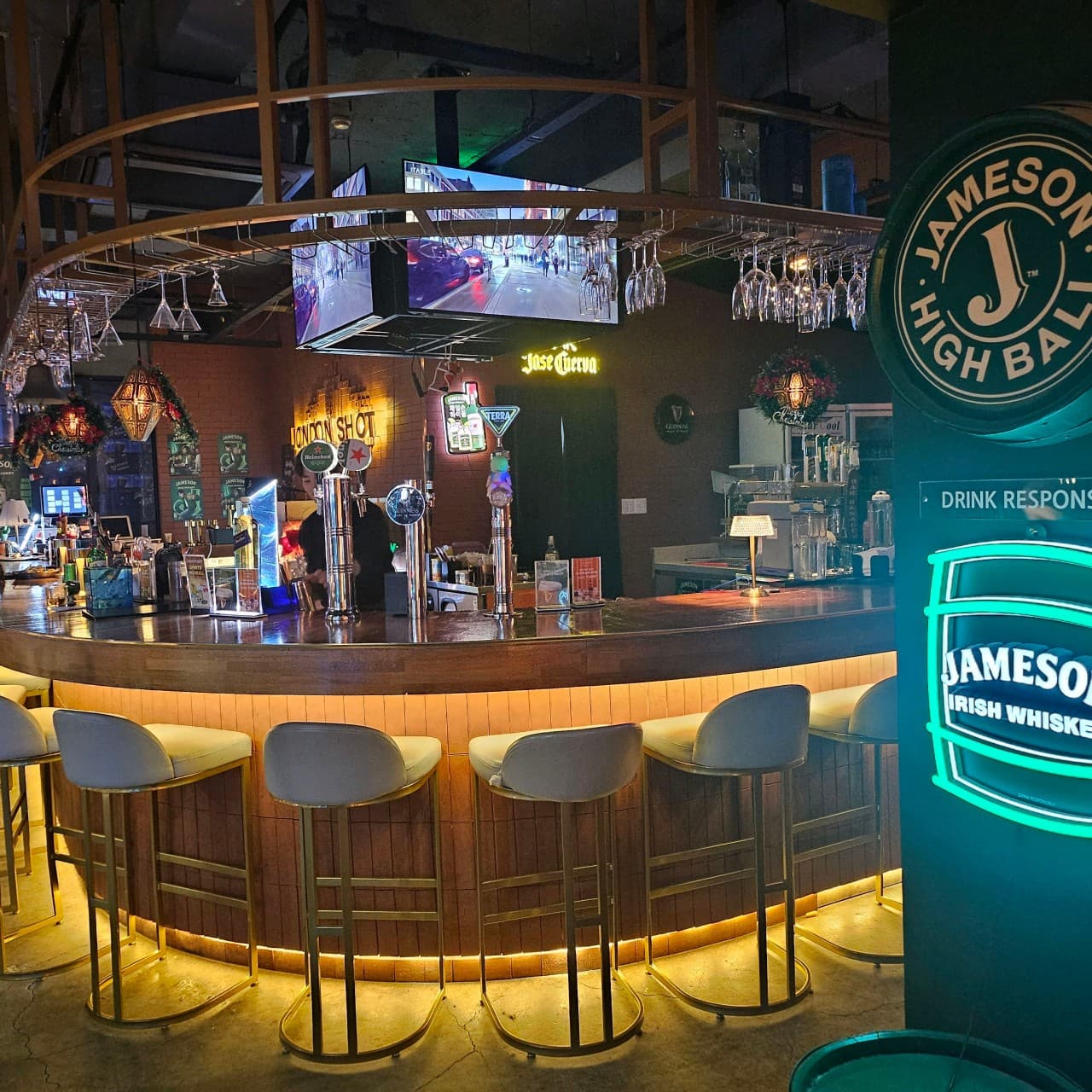 하단 조명이 비치는 원형 목재 카운터와 금색 프레임의 1인용 높은 의자들이 나란히 배치된 바 내부