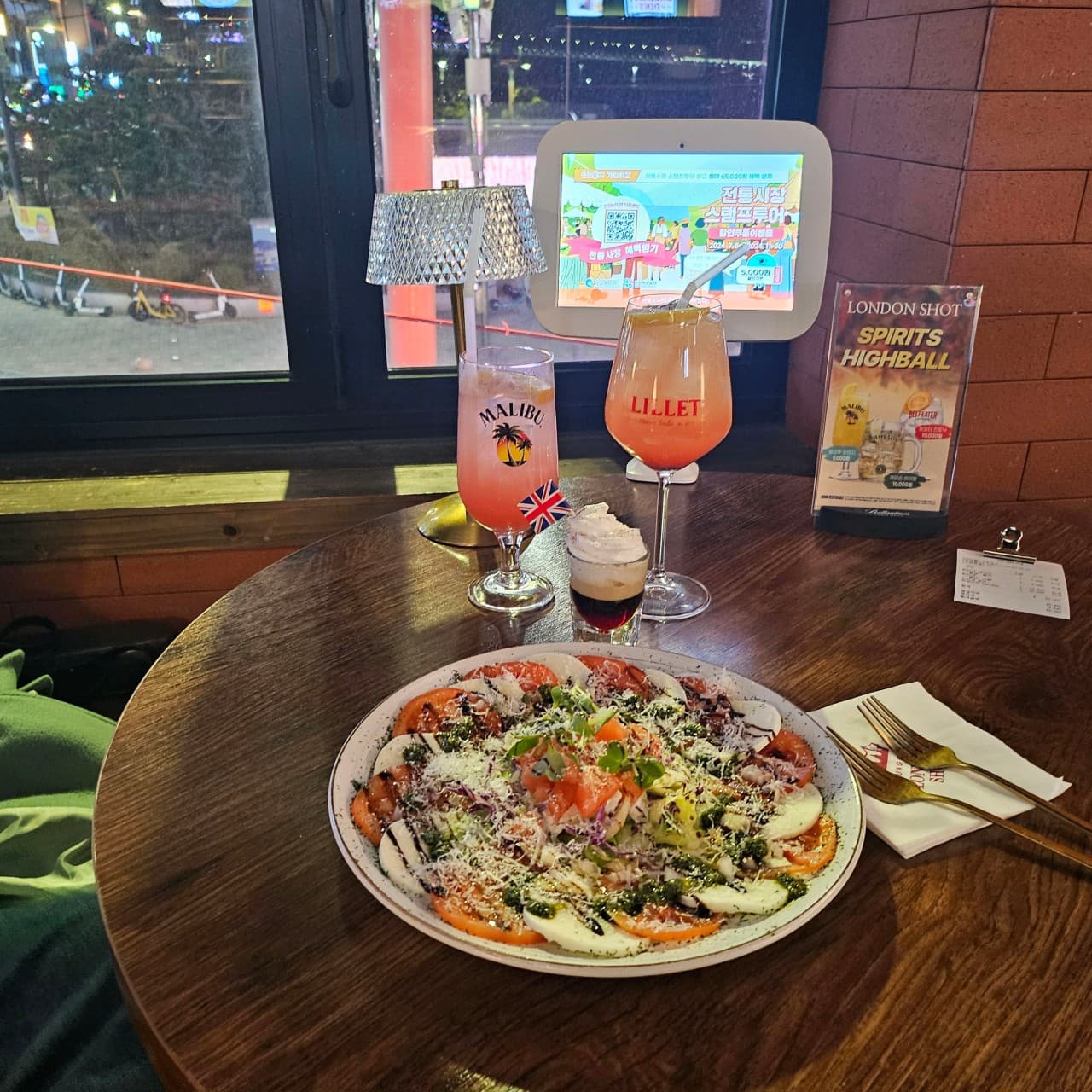 창가 원형 테이블 위 토마토 카프레제와 칵테일, 크림이 올라간 샷 잔과 은색 스탠드 조명이 놓인 좌석