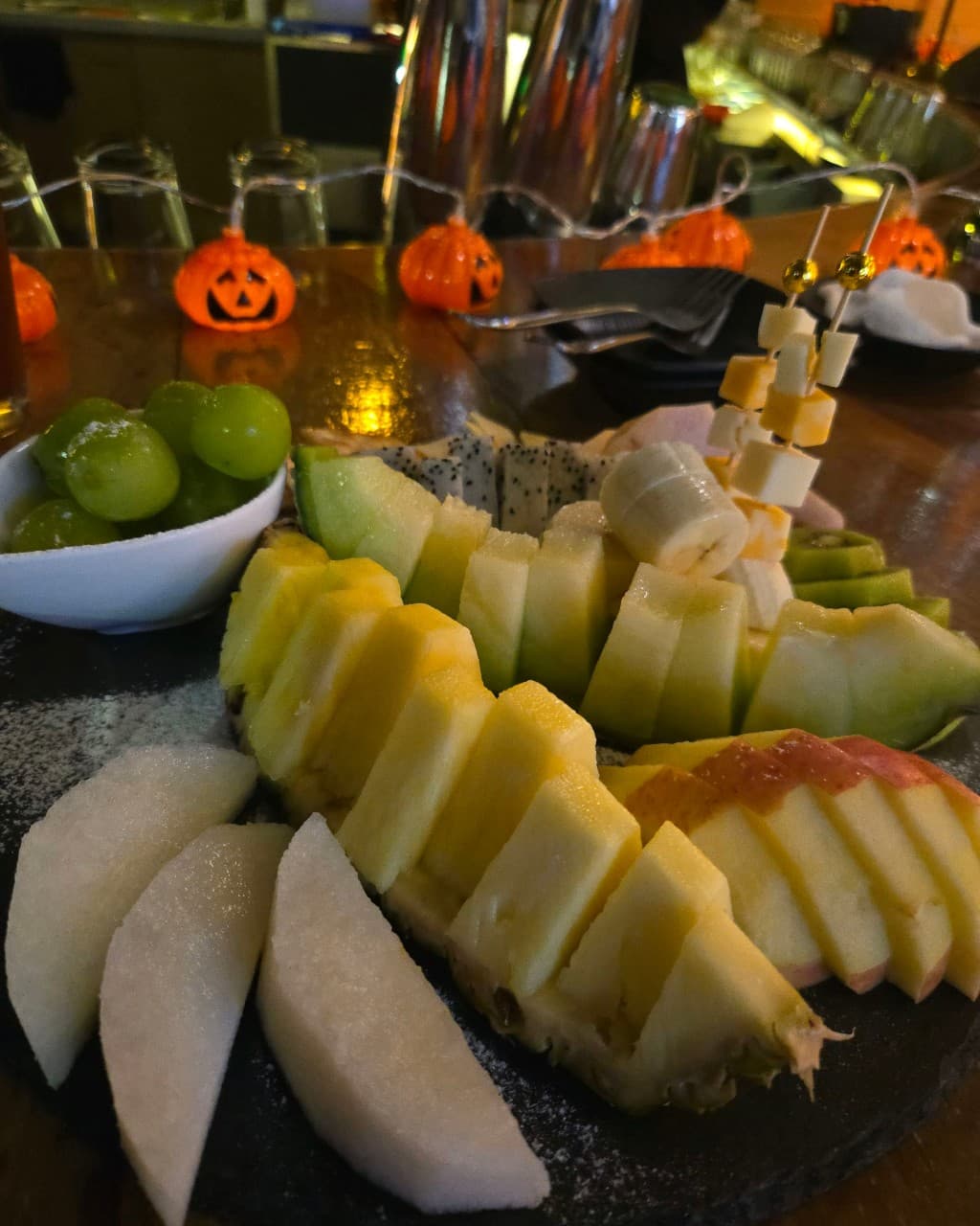 어두운 조명의 바 카운터 위 파인애플, 멜론, 사과 등 모둠 과일 접시와 호박 모양 전구 장식