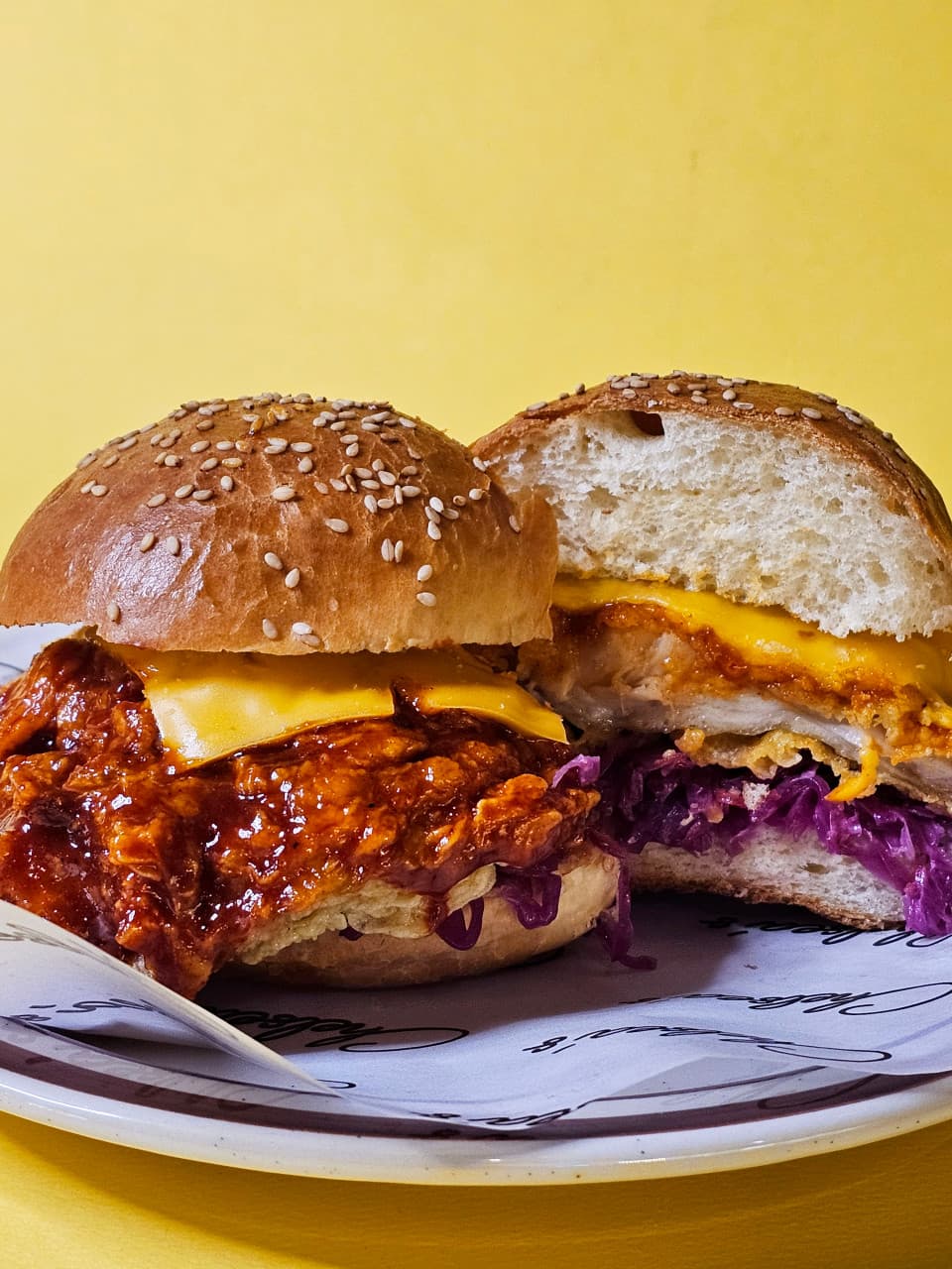 노란 배경 앞 접시에 놓인 참깨 번과 붉은 양념 패티, 녹은 치즈, 적채가 어우러진 버거 단면