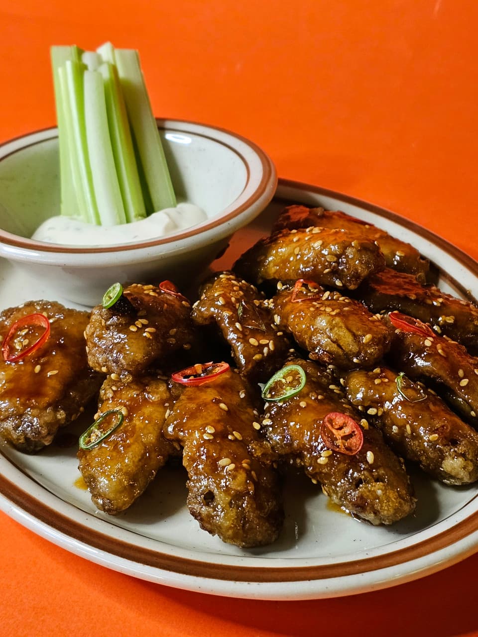 주황색 배경 위 소스가 발린 치킨 윙과 고추 고명, 흰 소스에 꽂힌 셀러리 스틱이 담긴 1인용 안주 플레이트