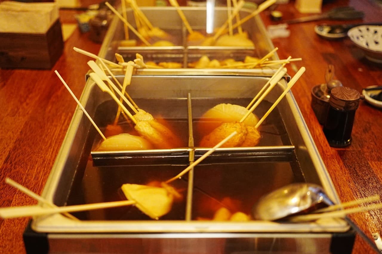 황색 조명이 비치는 목재 카운터 테이블 위 칸막이된 사각 용기에 담긴 다양한 오뎅 꼬치와 국자