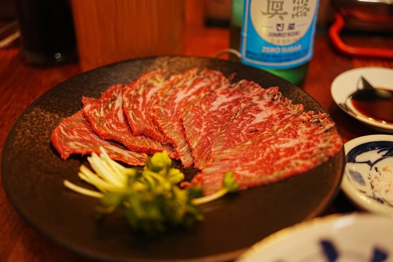 목재 테이블 위 얇게 썬 소고기와 와사비가 담긴 검은 접시, 소주병과 소스 종지가 놓인 1인 혼술 차림