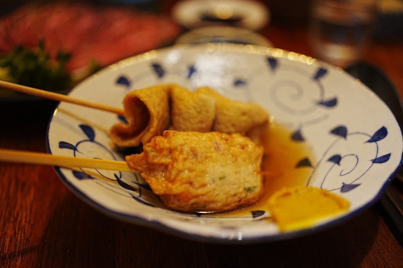 어두운 조명의 목재 카운터 위 푸른 문양 사발에 담긴 어묵 꼬치 두 개와 노란 겨자 소스