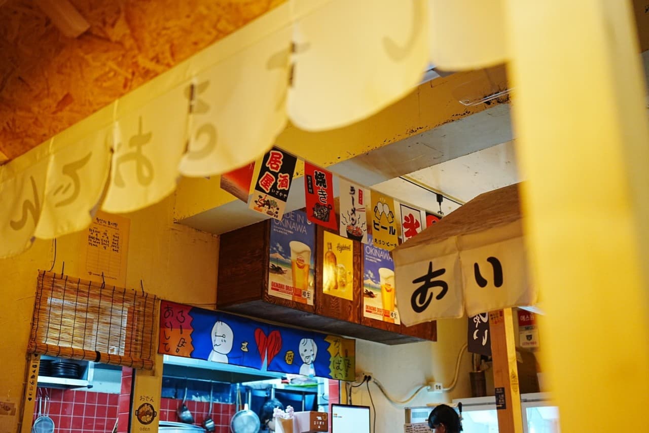 노란 조명 아래 일본어 문구가 적힌 가림막과 포스터가 부착된 내부 및 붉은 타일 벽면의 주방 전경