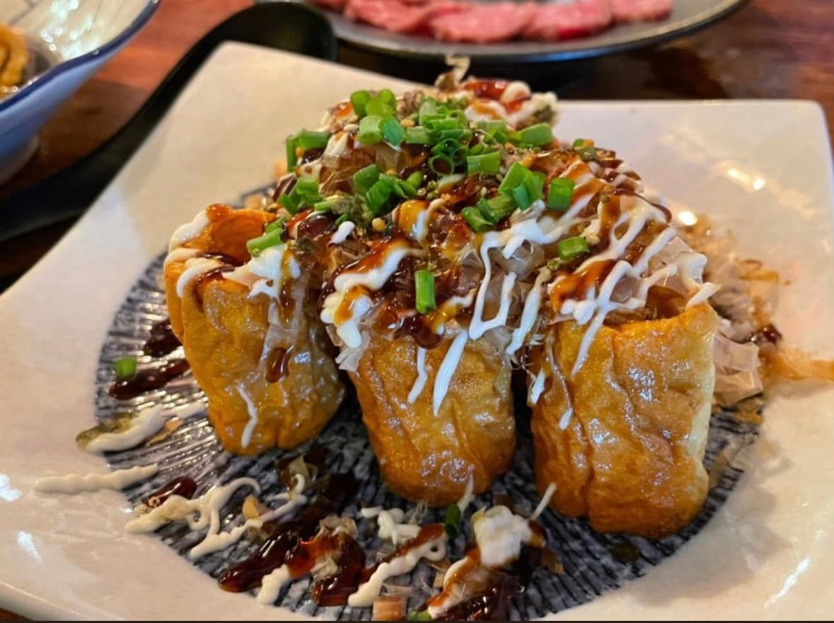 목재 카운터석 위 사각 접시에 담긴 유부 요리와 마요네즈, 데리야끼 소스, 가쓰오부시 및 쪽파 고명