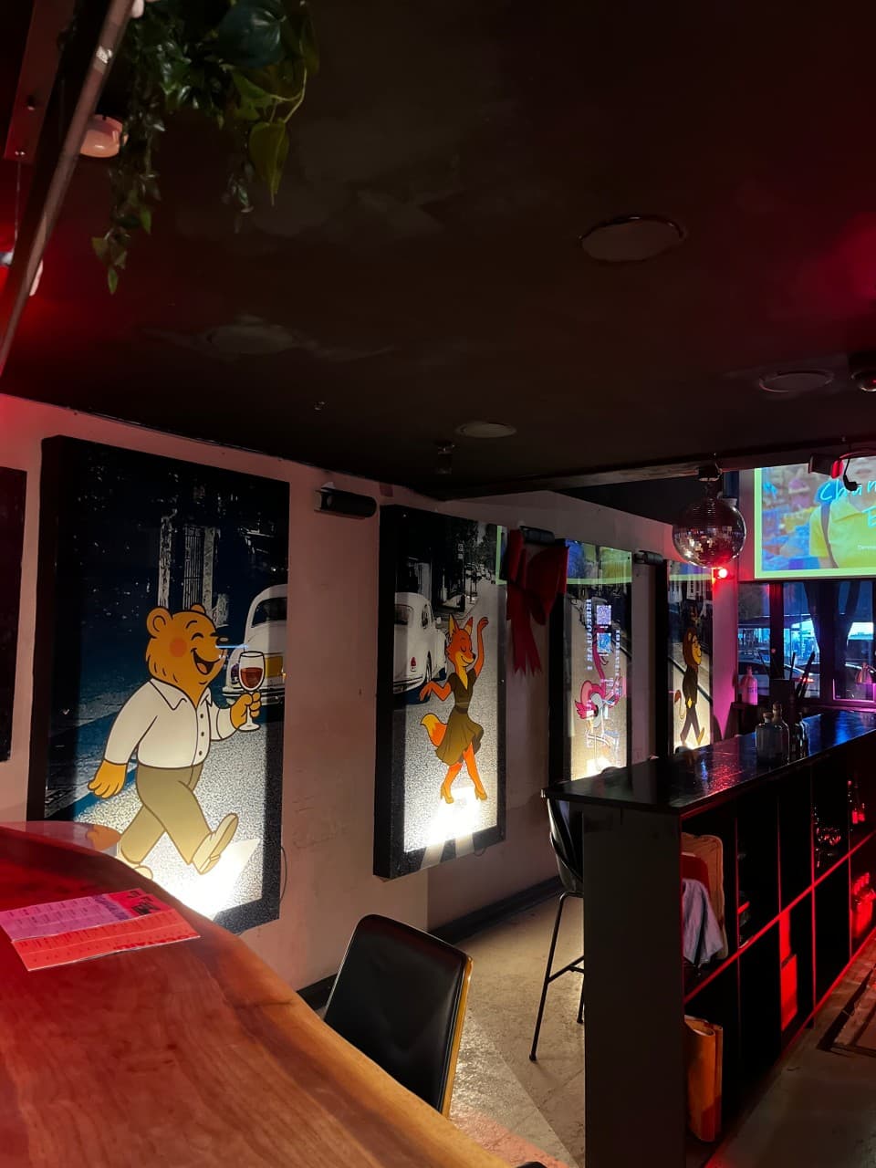 붉은 조명이 비치는 어두운 실내에 나무 카운터와 1인용 바 좌석이 마련되어 있고 벽면에는 와인 잔을 든 캐릭터 조명 액자가 설치된 공간