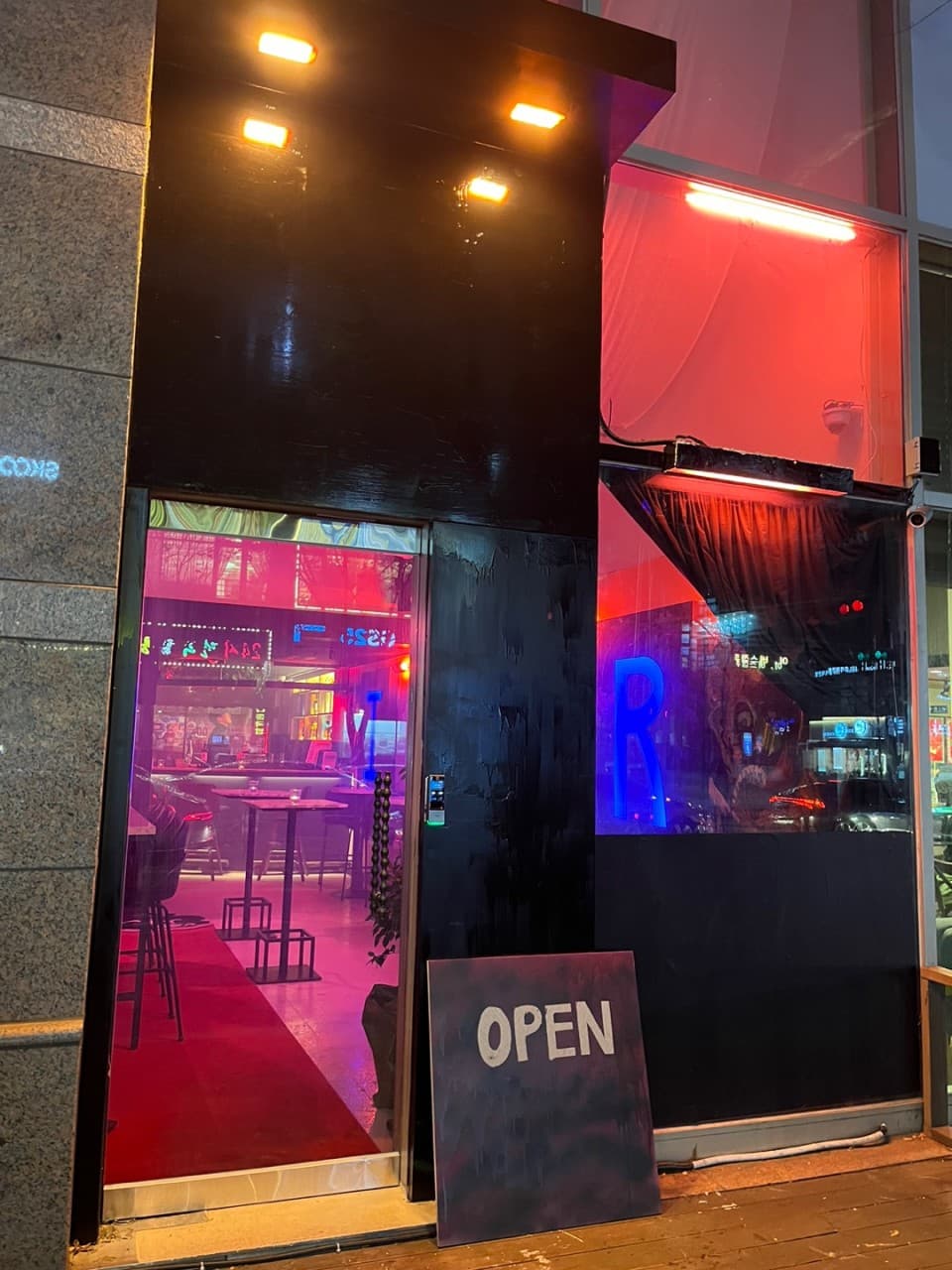 붉은 조명과 푸른 네온사인이 비치는 검은색 매장 입구. 내부에는 높은 바 테이블과 1인용 의자가 배치되어 있는 모습