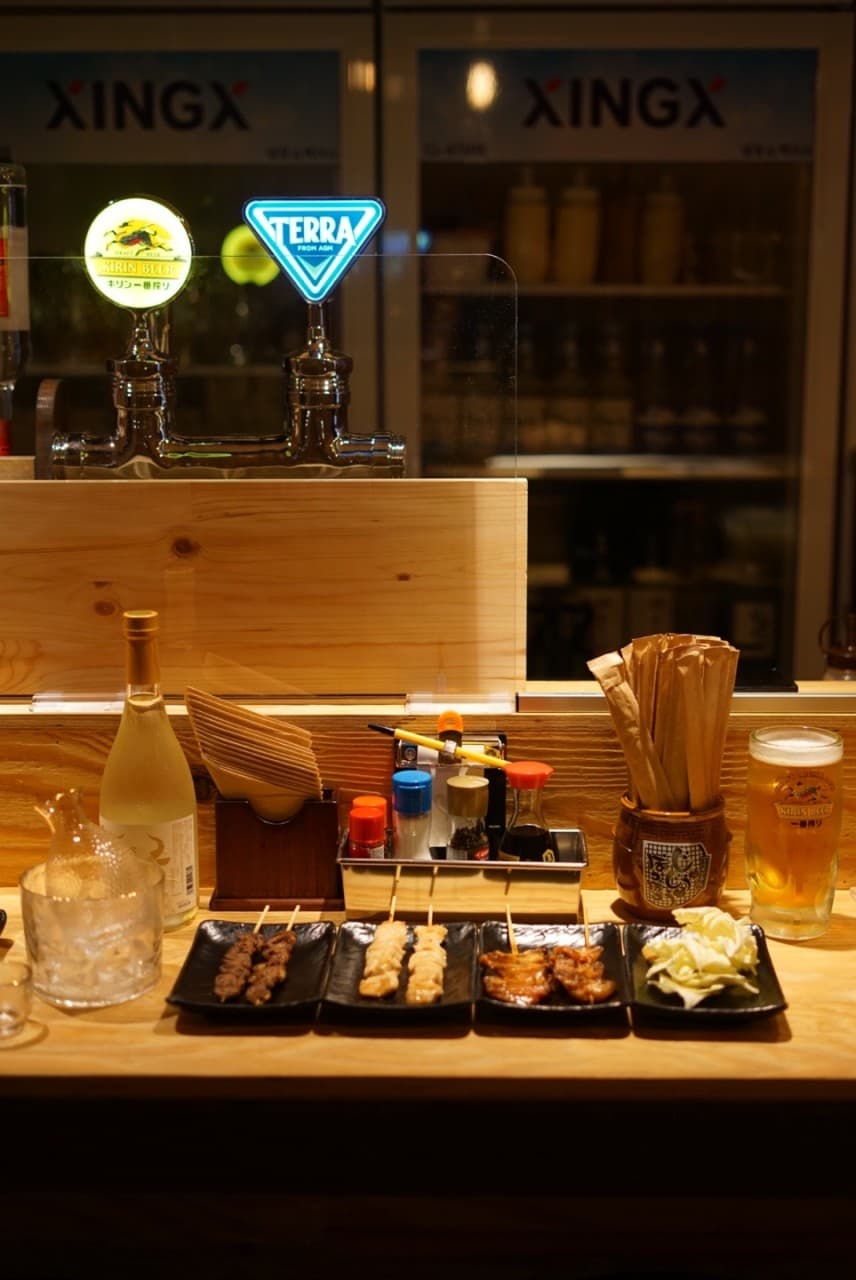 목재 카운터 위 네 종류의 꼬치구이와 양배추, 생맥주 잔과 사케 병이 나란히 놓인 1인용 좌석 전경