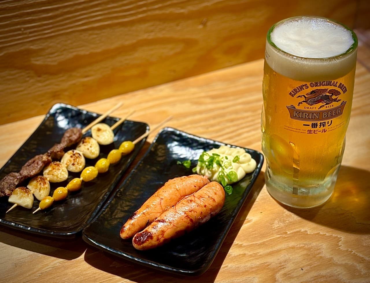 나무 질감의 테이블 위 생맥주 한 잔과 명란구이, 세 종류의 꼬치구이가 담긴 검은색 접시