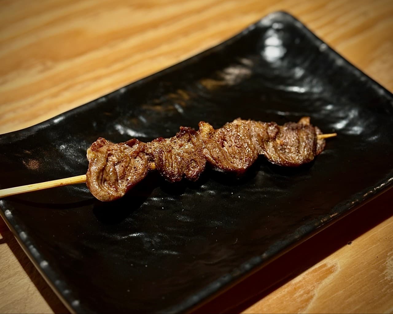 나무 질감이 드러나는 바 테이블 위 검은색 사각 접시에 담긴 숯불 고기 꼬치 구이 한 점