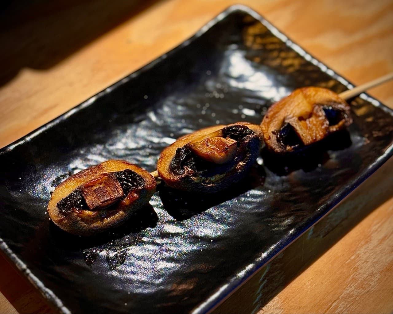 노란 조명이 비치는 나무 카운터 위 검정색 사각 접시에 담긴 버섯 꼬치구이와 목재 테이블의 질감