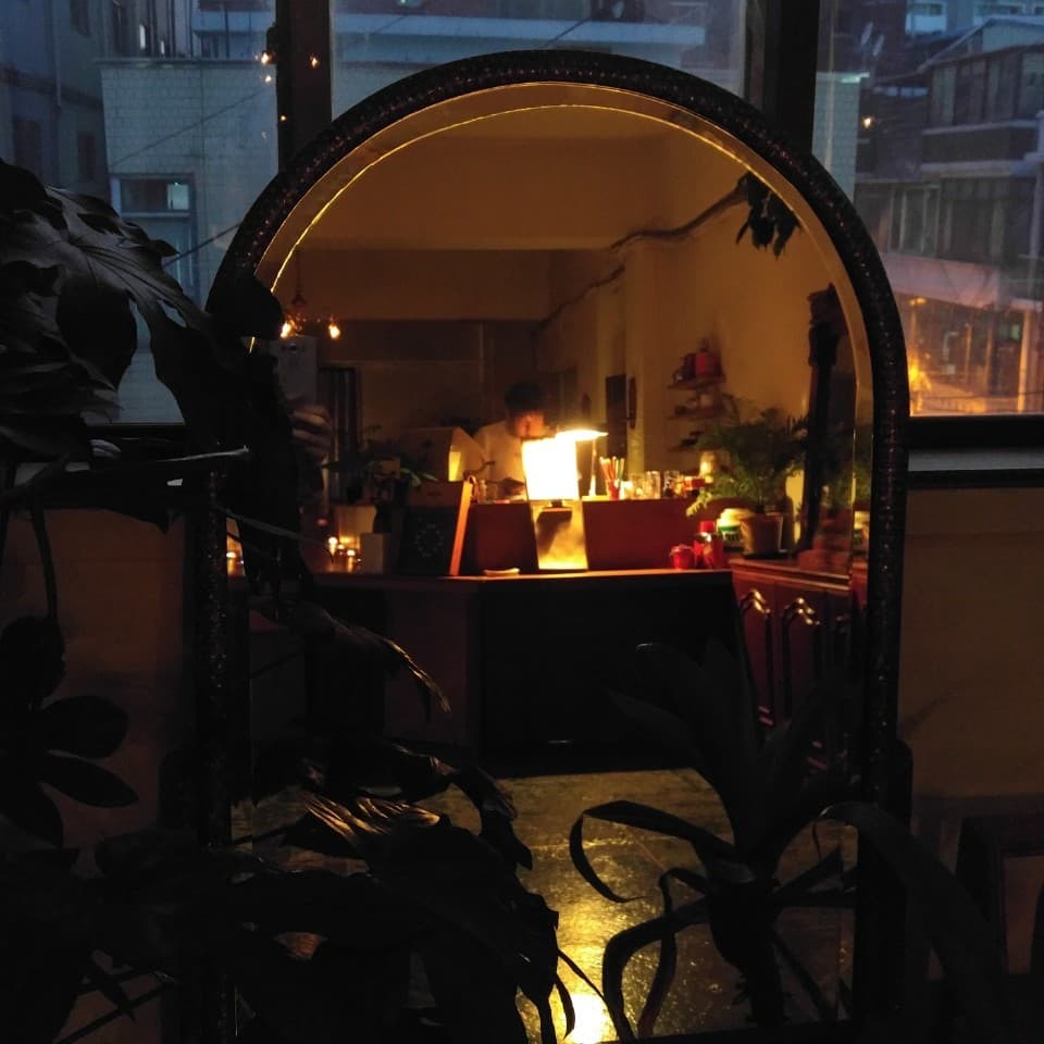 어두운 실내의 식물 실루엣 너머 아치형 거울에 비친 주황색 조명의 바 카운터와 1인용 좌석이 배치된 전경