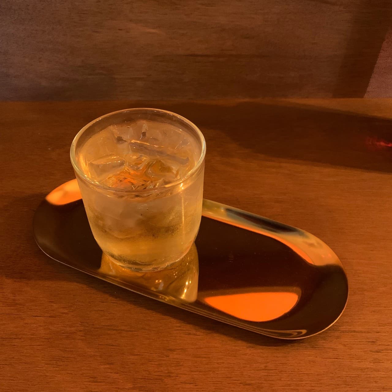 어두운 나무 질감의 카운터 위 금색 트레이와 얼음이 담긴 하이볼 잔에 반사된 주황색 조명
