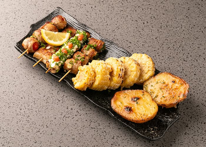 검은색 접시 위 토마토 고기말이와 쪽파를 얹은 꼬치, 구운 옥수수 및 감자로 구성된 1인용 모둠 안주