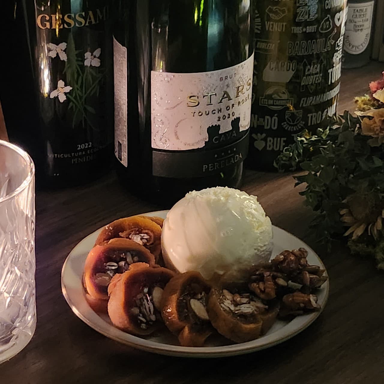 어두운 목재 카운터 위 아이스크림과 곶감 말이 접시, 투명한 크리스털 잔 뒤로 나열된 와인 병들