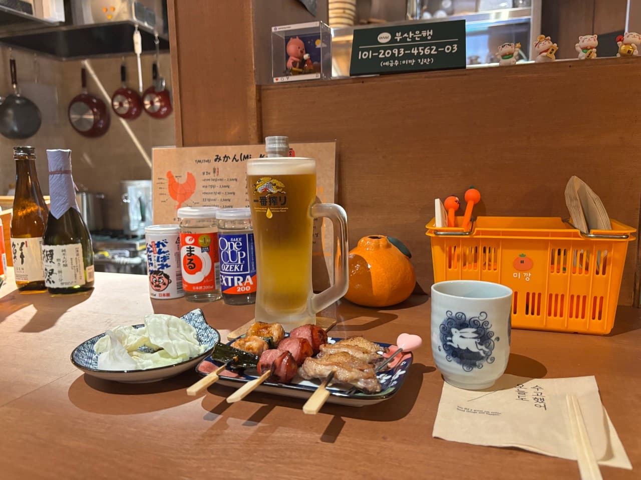 갈색 나무 바 테이블 위 생맥주 잔과 꼬치구이 접시, 양배추 샐러드와 사케병이 차려진 1인용 좌석 전경