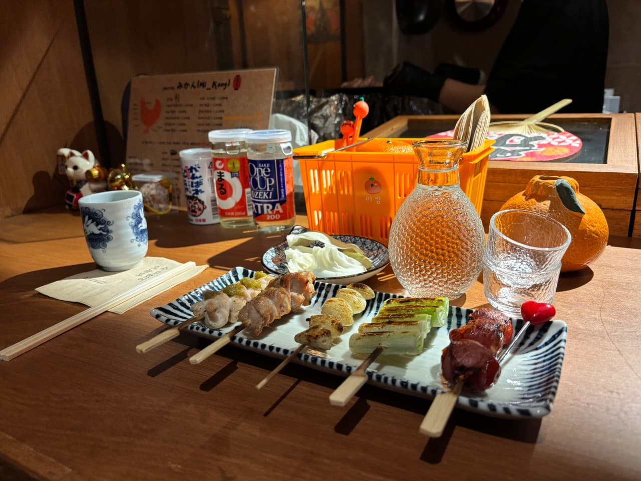 황색 조명이 비치는 나무 바 테이블 1인석에 차려진 닭꼬치 구이 접시와 투명한 유리 술병 및 작은 잔