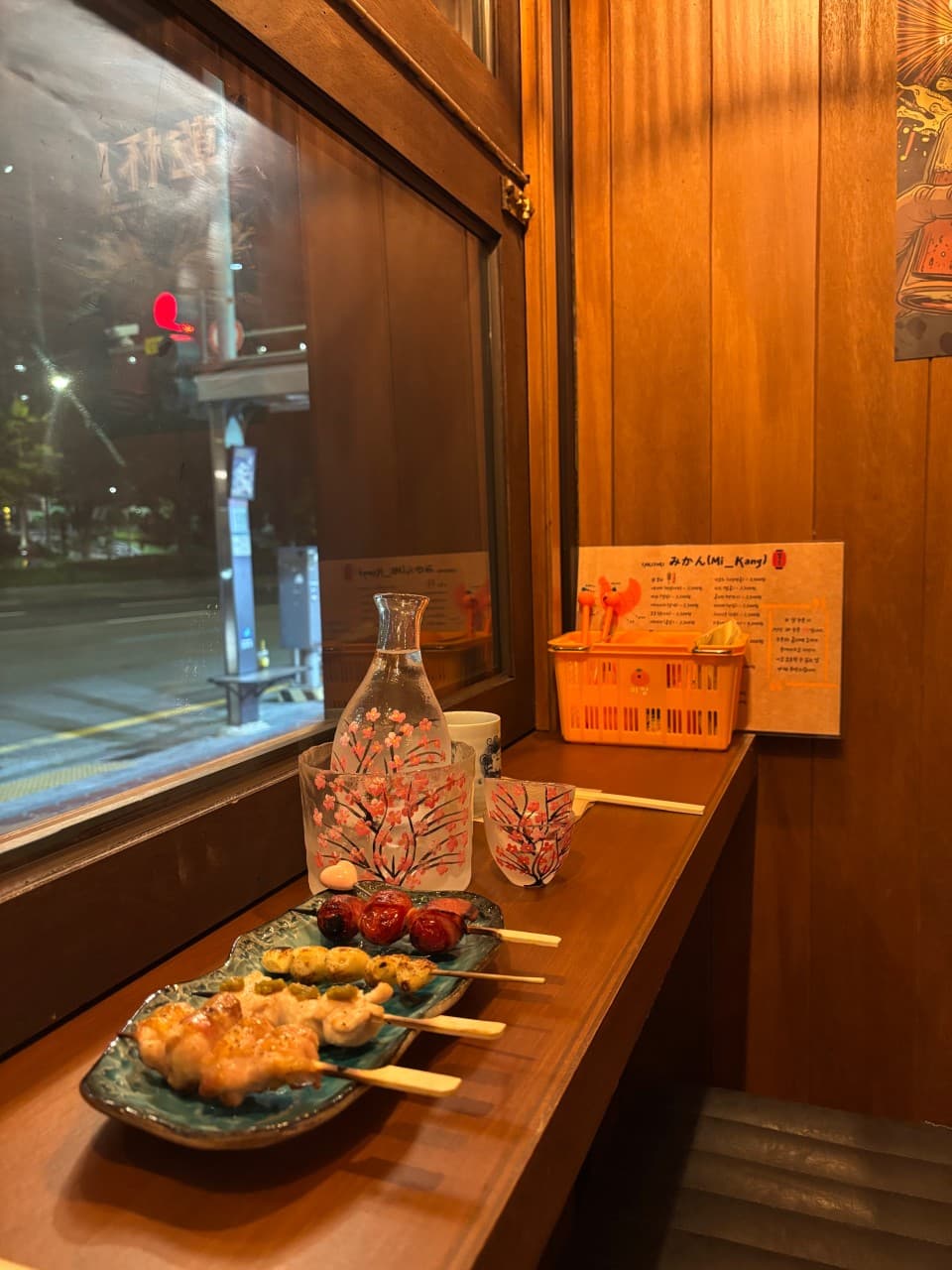 밤거리가 보이는 창가 목재 카운터석에 차려진 닭꼬치 구이 접시와 벚꽃 문양 사케병 및 전용 술잔