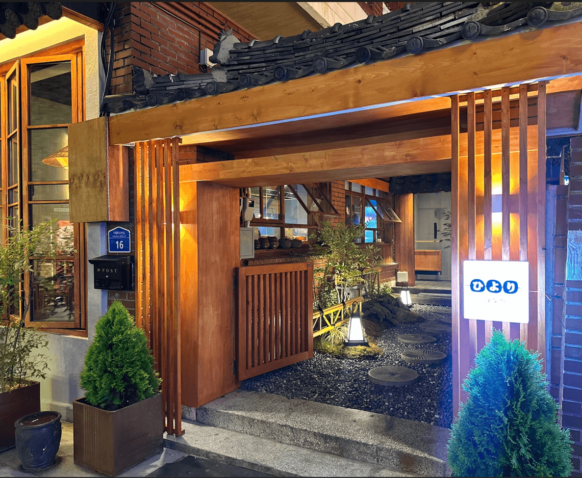 기와를 얹은 목재 대문 안쪽으로 자갈과 디딤돌이 깔린 마당이 보이며 노란 조명 아래 좁은 통로가 이어지는 술집 입구 전경