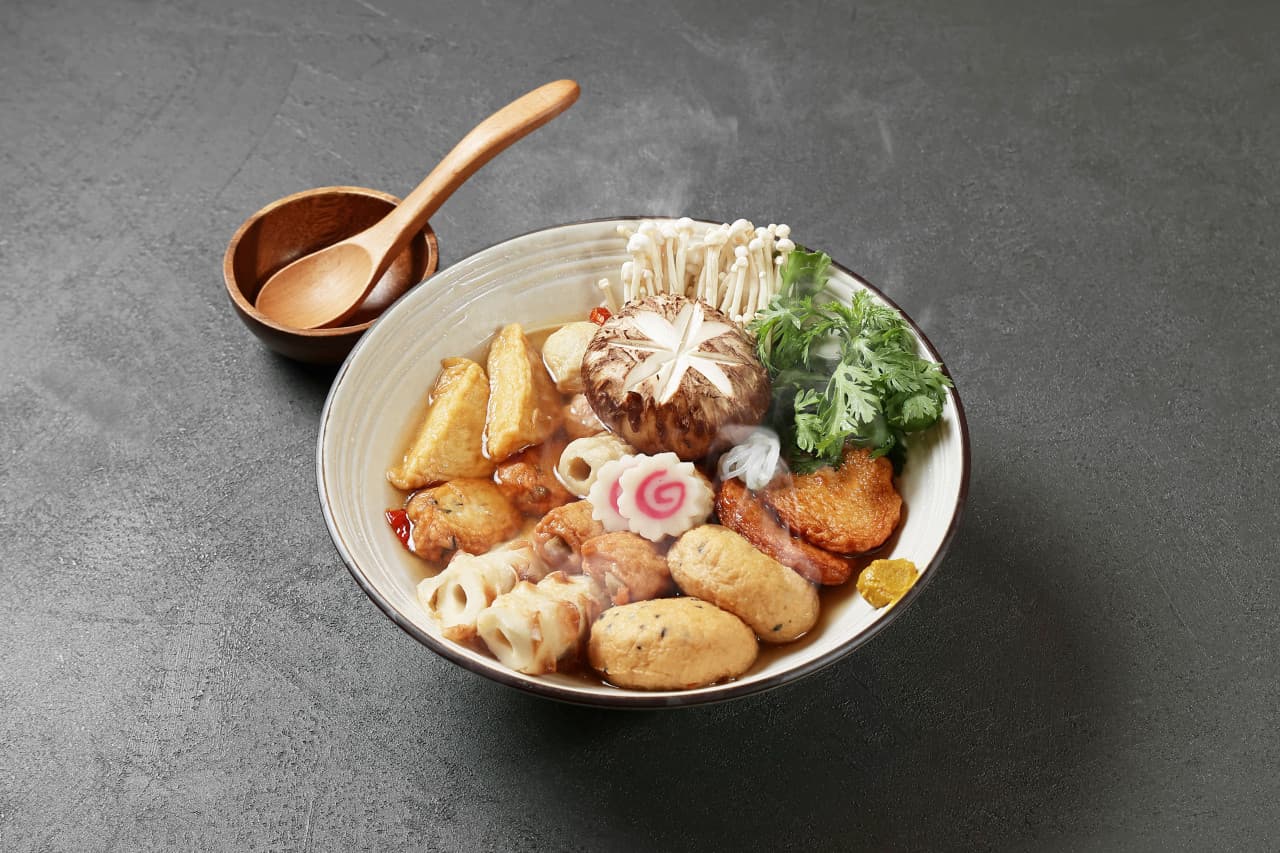 어두운 무채색 테이블 위 김이 피어오르는 1인용 어묵탕 한 그릇과 나무 소재의 작은 수저 세트