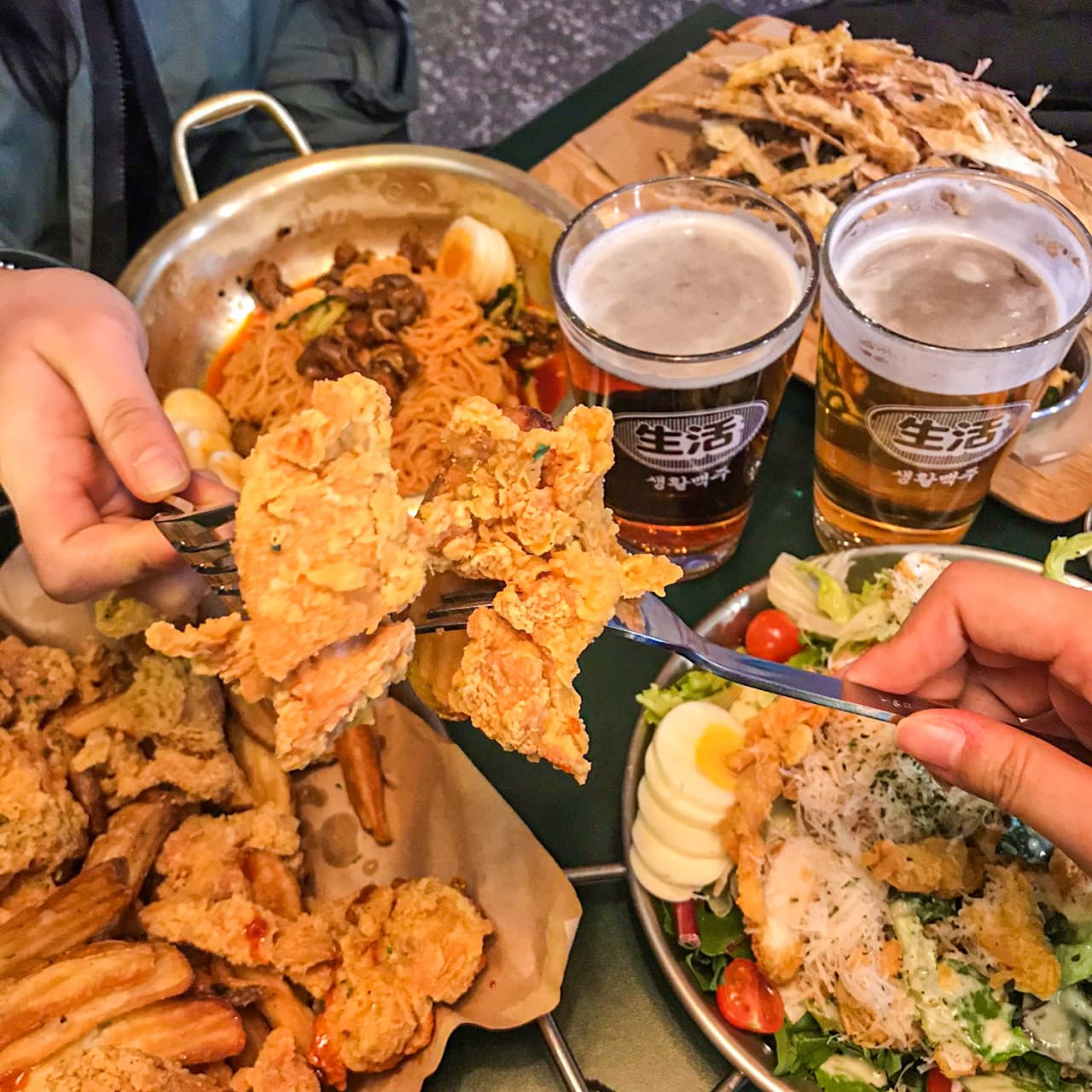 포크로 집어 올린 프라이드 치킨과 거품이 있는 맥주, 비빔면과 샐러드 등 안주가 차려진 테이블