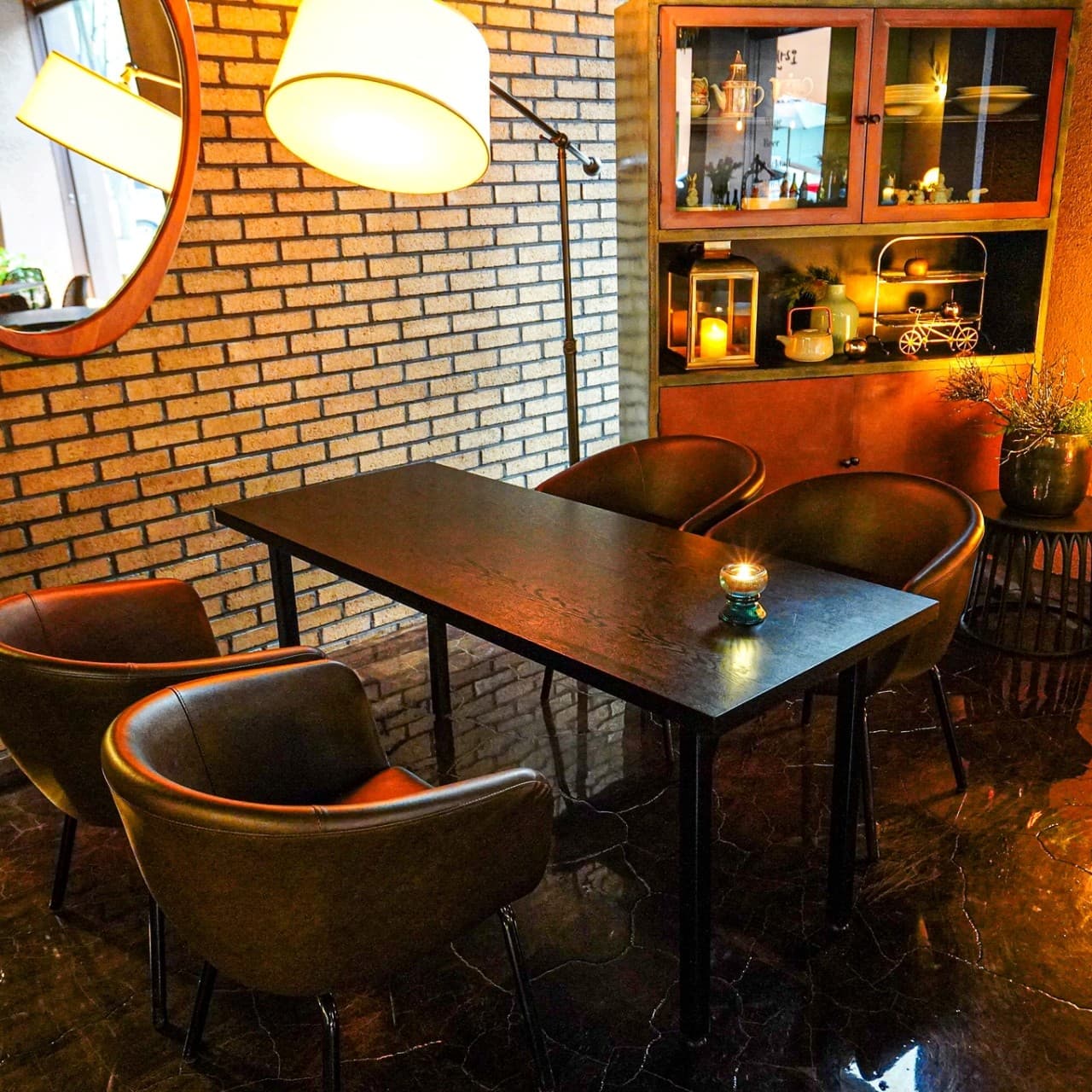 노란 조명이 켜진 스탠드 아래 검은색 테이블과 1인용 가죽 의자, 벽돌 벽과 장식장이 배치된 실내 전경
