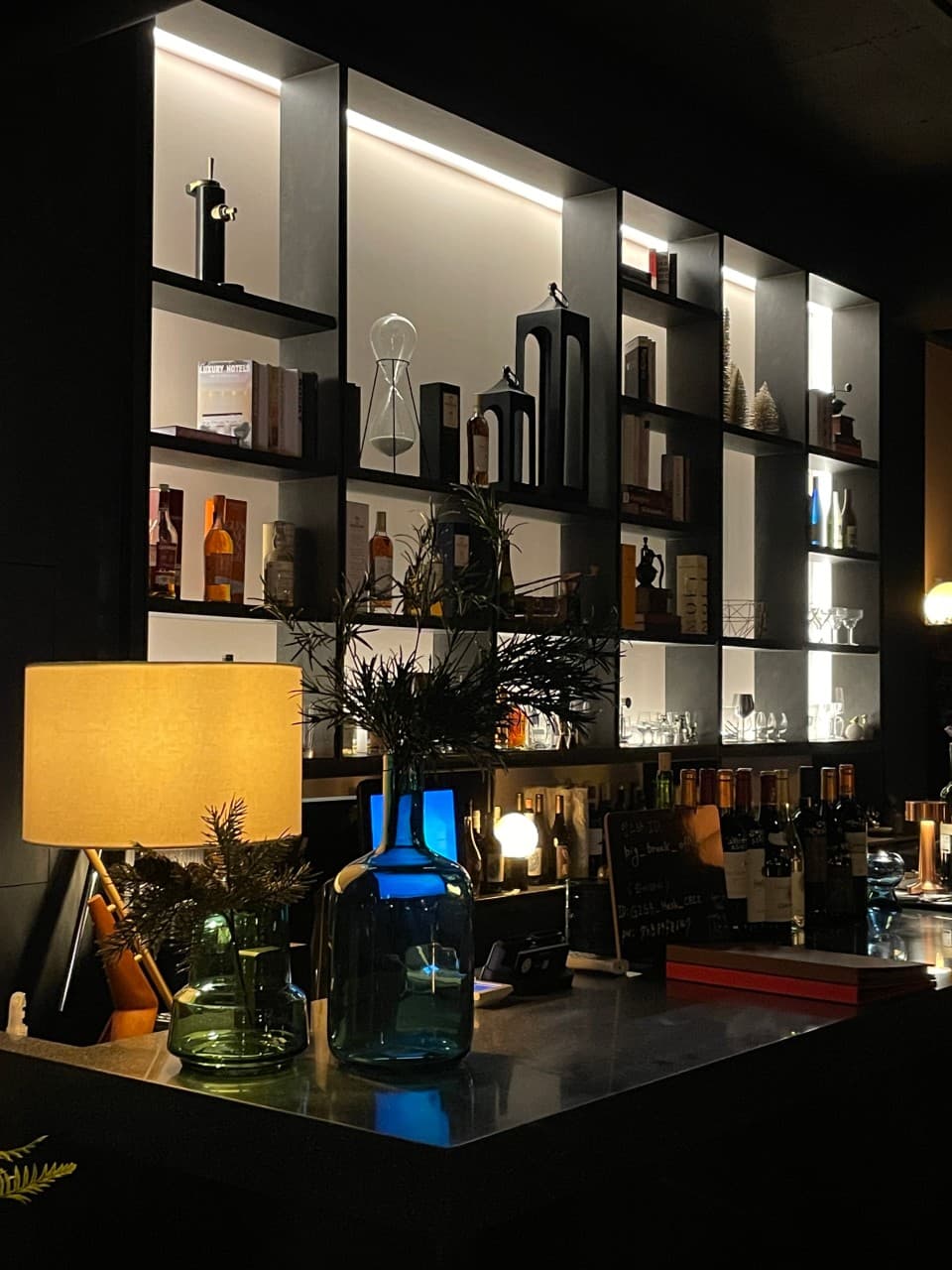 백라이트가 켜진 대형 진열장 앞 바 카운터에 놓인 노란 전등과 푸른 유리병, 여러 종류의 와인병이 나열된 어두운 실내