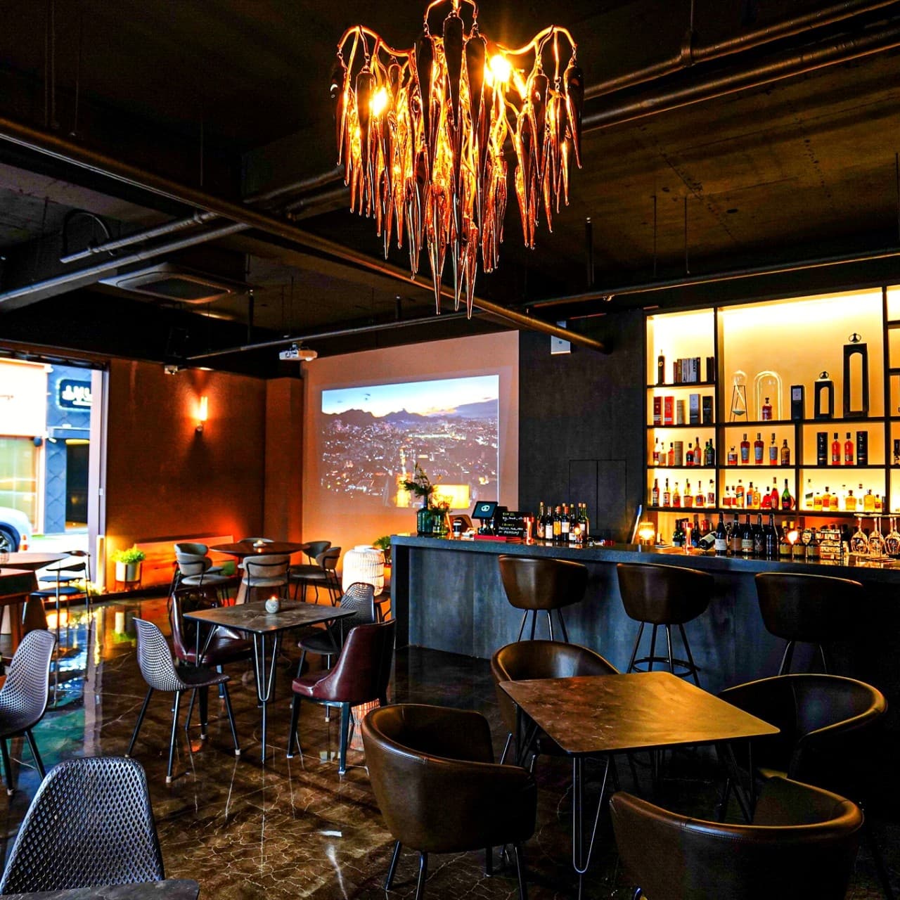 금색 샹들리에 아래 긴 바 카운터와 높은 의자가 배치된 바 내부. 벽면 진열장에는 다양한 술병이 있고 대형 스크린에서 영상이 나옴.