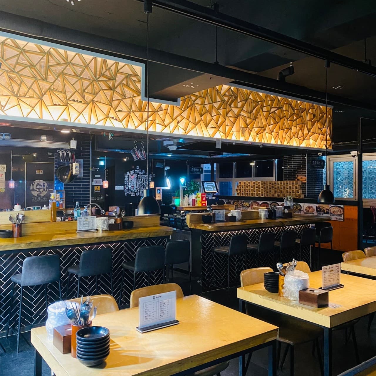 기하학적 무늬의 상단 조명 아래 바 테이블과 높은 의자가 늘어선 실내. 나무 식탁 위에는 검은 식기류와 메뉴판이 비치되어 있는 모습.