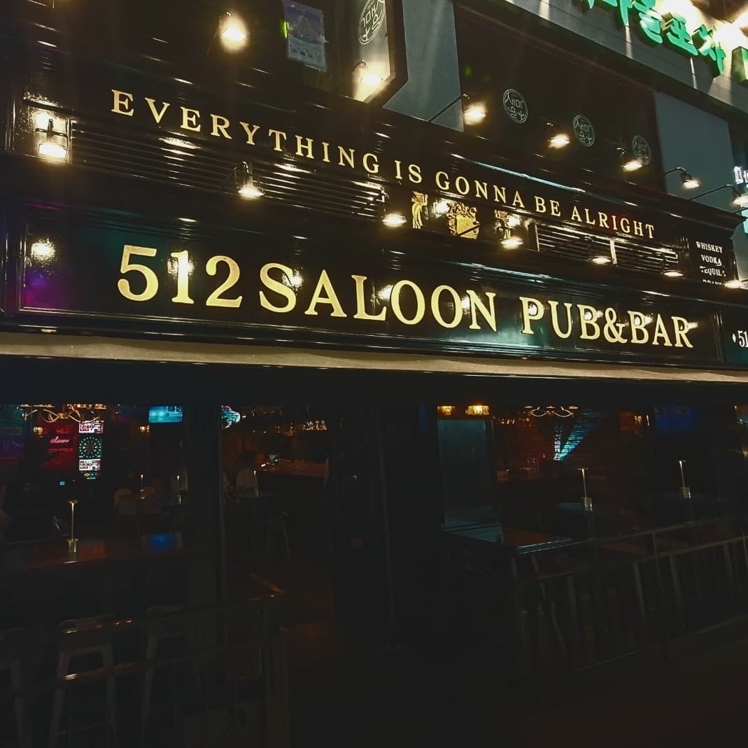 어두운 외벽에 노란 조명이 켜진 금색 영문 간판과 창가를 따라 나란히 배치된 1인용 바 테이블 및 의자