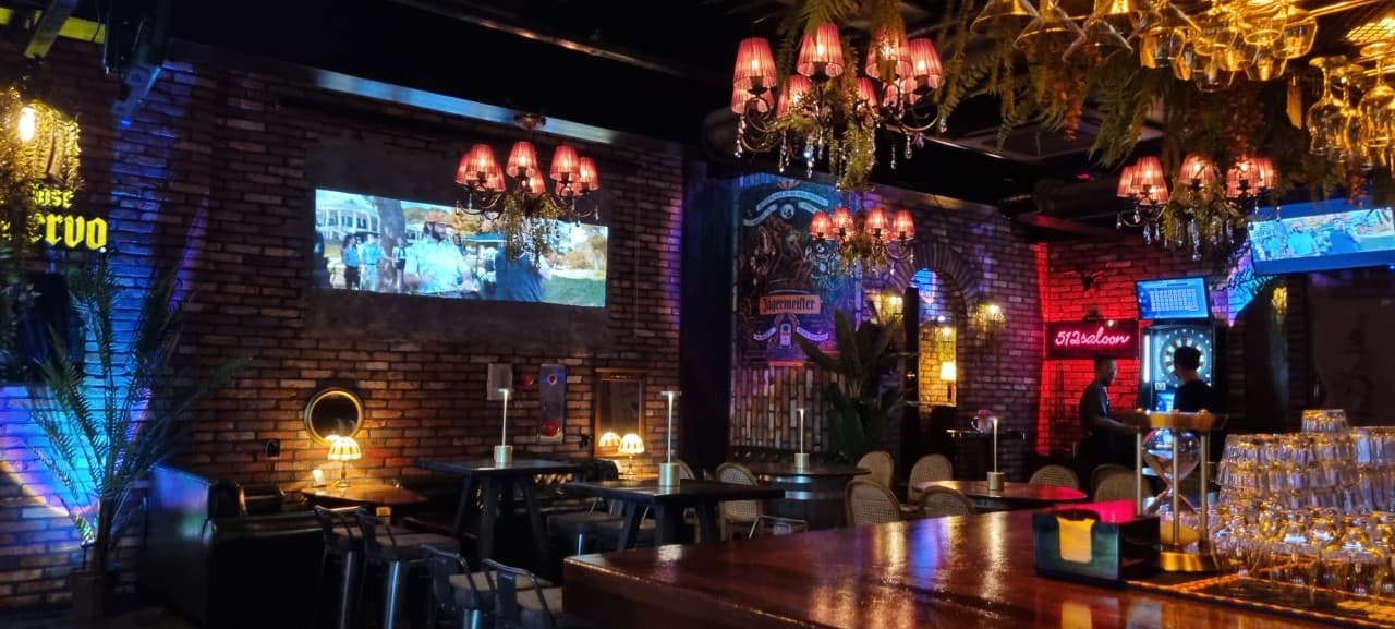 붉은 샹들리에 조명 아래 벽돌 벽면의 스크린과 바 카운터, 1인용 의자가 배치된 테이블 전경