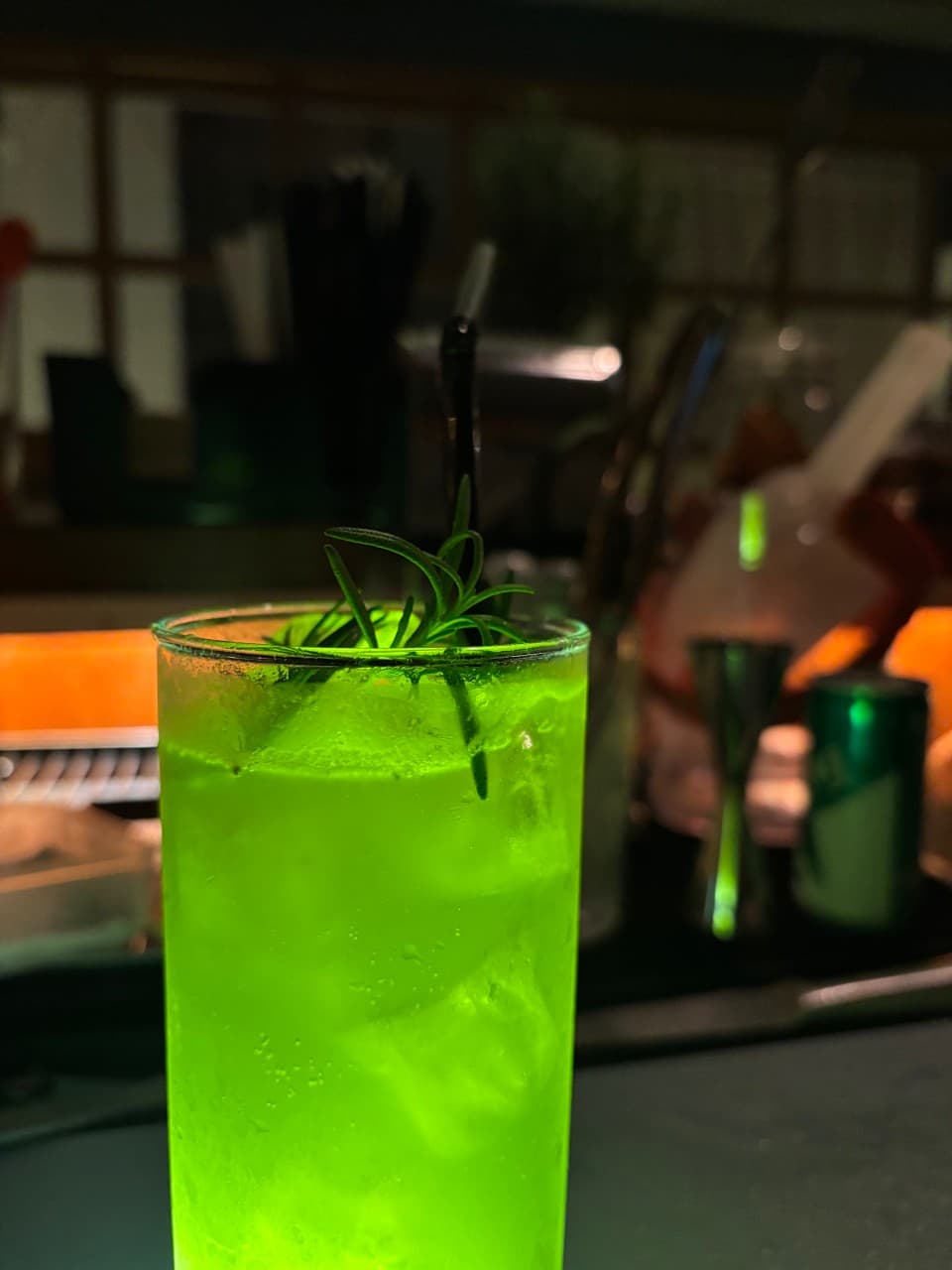 어두운 바 테이블 위에 놓인 로즈마리가 꽂힌 연두색 하이볼 잔과 뒤로 보이는 주황색 조명