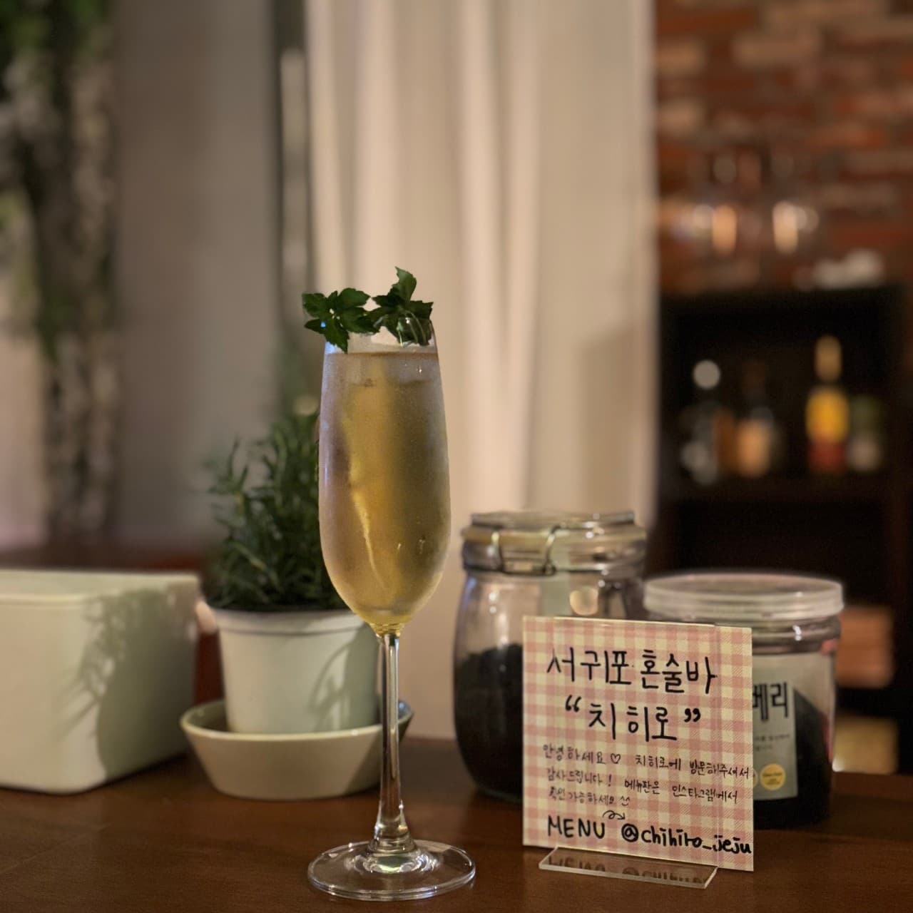 어두운 실내 목재 카운터 위 허브 장식 칵테일 잔과 작은 화분 및 혼술 안내 카드