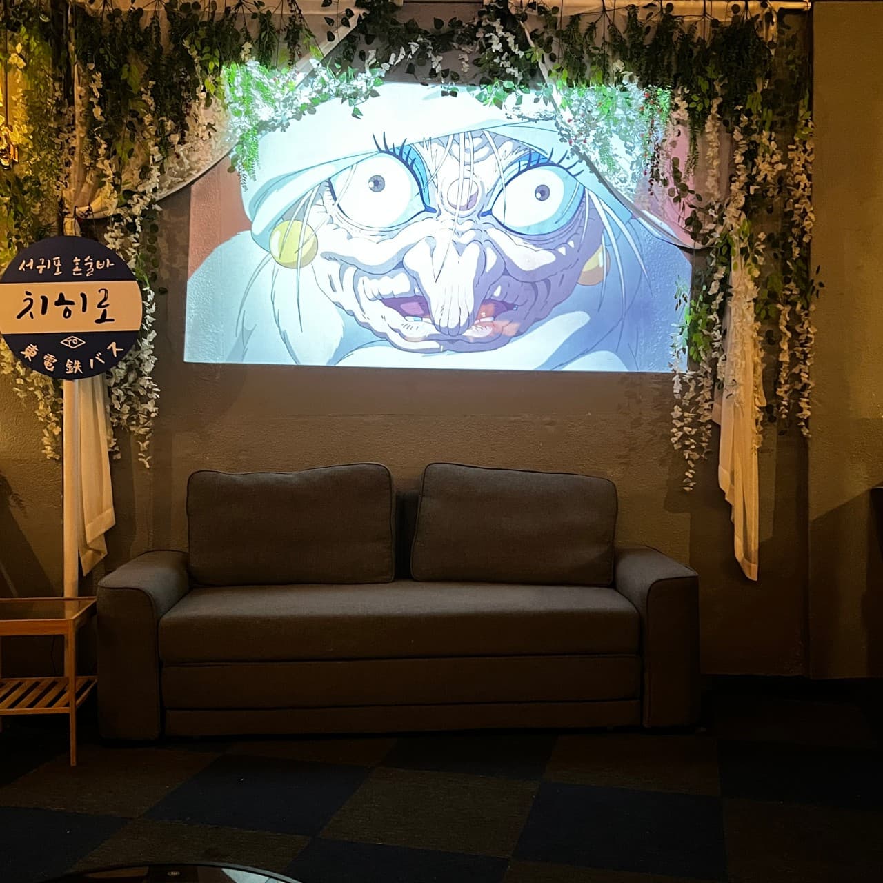 어두운 조명 아래 애니메이션 영상이 상영되는 벽면과 조화 넝쿨로 장식된 회색 소파 좌석