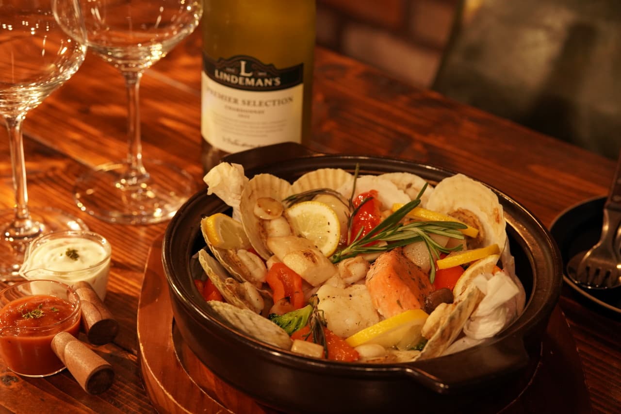 나무 질감의 카운터 위 가리비와 연어가 담긴 냄비 요리, 화이트 와인병과 와인 잔 및 작은 소스 용기