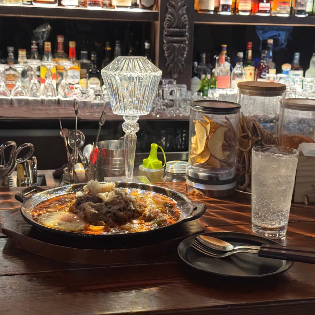 어두운 목재 바 테이블 위 1인용 무쇠 팬 요리와 하이볼 잔, 크리스탈 스탠드 조명