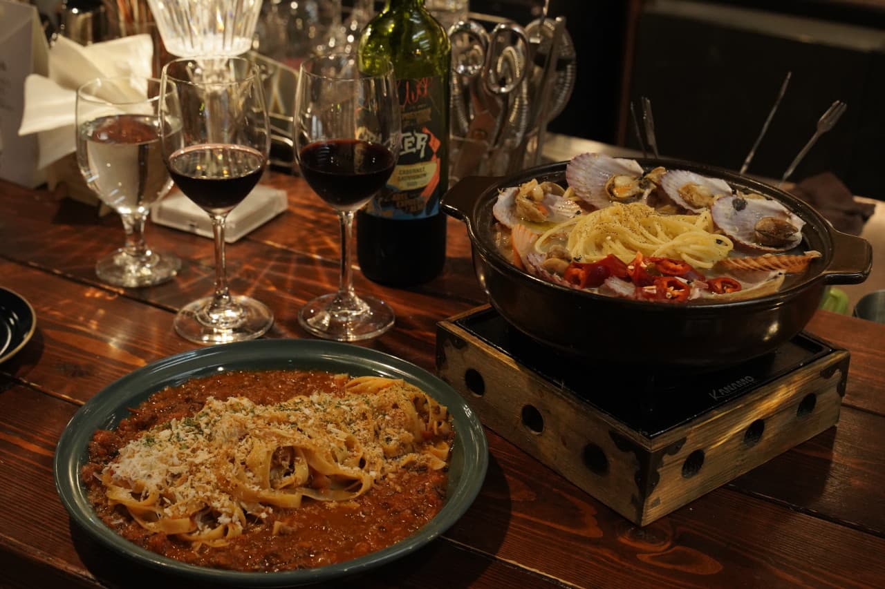 어두운 조명의 나무 카운터 석에 놓인 라구 파스타와 가리비 해산물 전골, 레드 와인 병과 잔